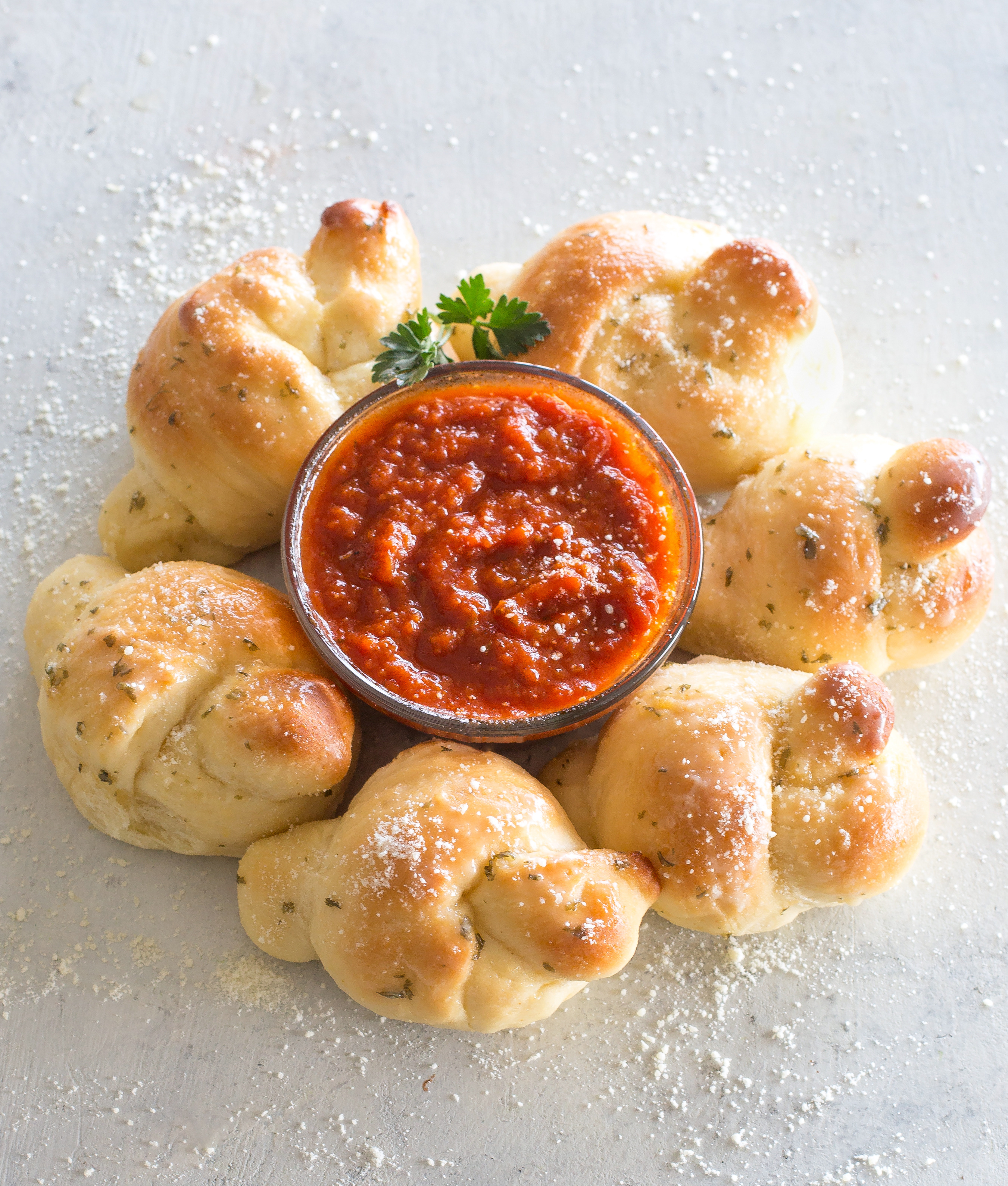Soft homemade garlic knots brushed with garlic butter and Parmesan next to a bowl of marinara